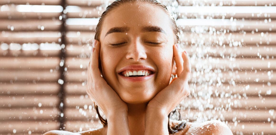 Mulher sorridente de olhos fechados desfrutando de um banho relaxante sob um chuveiro moderno, com água escorrendo pelo rosto e ombros em um banheiro iluminado.